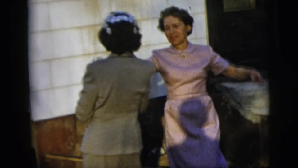 A woman in a lavender dress faces the camera mid-dance while the woman in a gray suit is seen from behind near the building exterior.