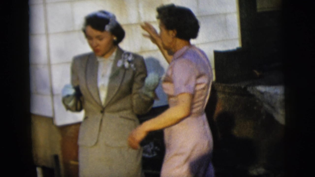 Two women in dressy clothing stand close together beside a shingled wall.