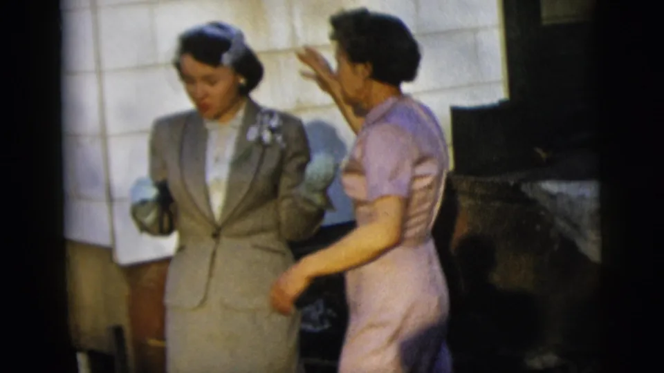 Two women in 1950s attire stand near a shingled wall in New York City home-movie footage, one in a grey suit and one in a pale purple dress.