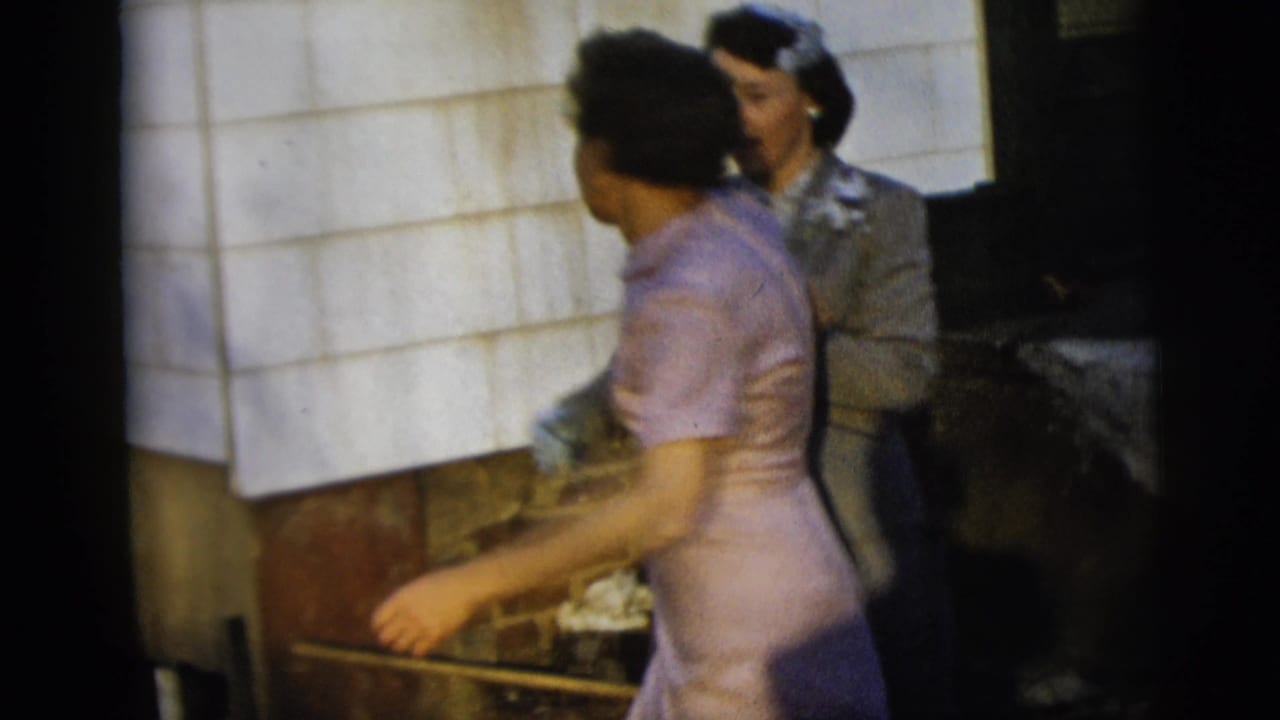 A woman in a pale purple dress moves past a woman in a grey suit during a brief 1958 New York City home-movie dance moment.