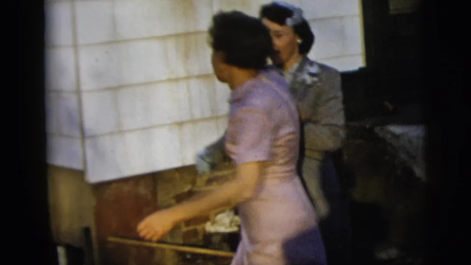 A woman in a pale purple dress moves past a woman in a grey suit during a brief 1958 New York City home-movie dance moment.