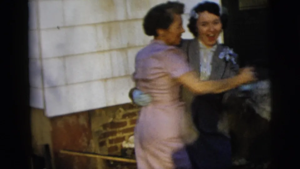 Two women smile and embrace in a close dance near a shingled wall, captured in grainy 1958 New York City 8mm footage.