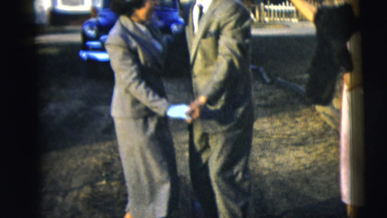 Two adults in suits hold hands while dancing on a lawn with a blue car behind them.