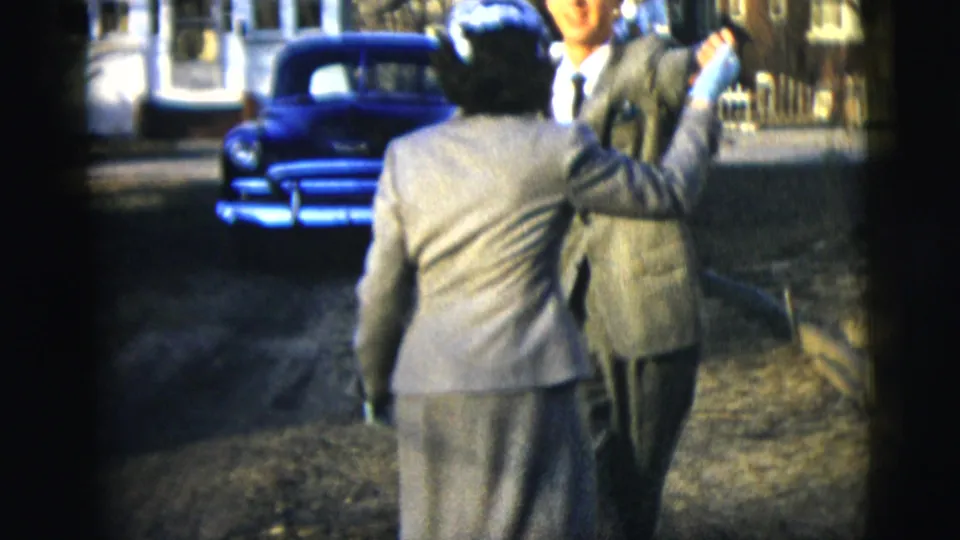 A woman in a light suit turns with her arm raised while dancing with a suited man on a New York lawn in 1958 home-movie footage.
