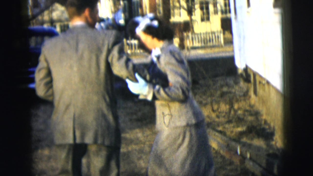 A suited man and gloved woman stand close together on a driveway.