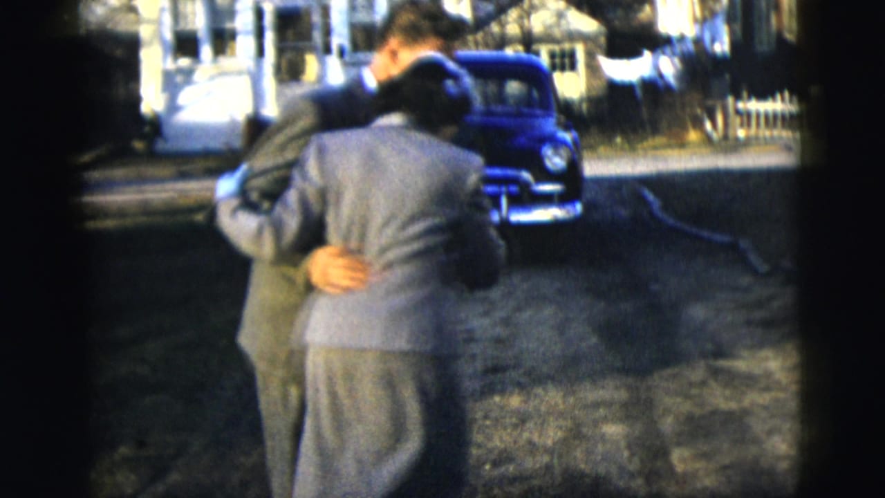 Couple dances in the street with a blue car and houses behind them.