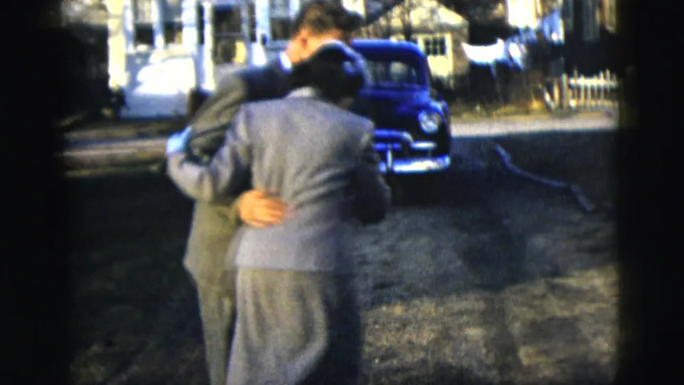 A 1958 New York City home-movie frame shows the couple embracing while dancing near a blue mid-century car on a residential street.