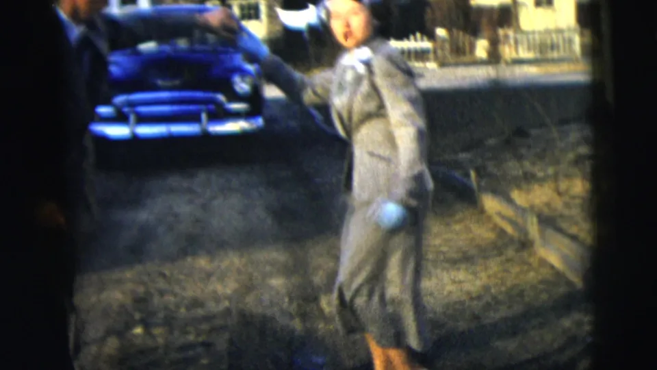 The woman in a gray suit and white heels turns during the dance, with a blue car and residential street setting behind her.