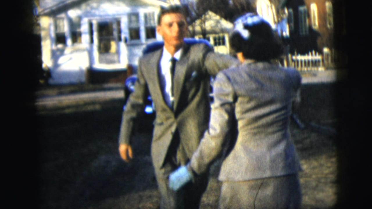 Suited man and woman dance or turn toward each other on a residential street.