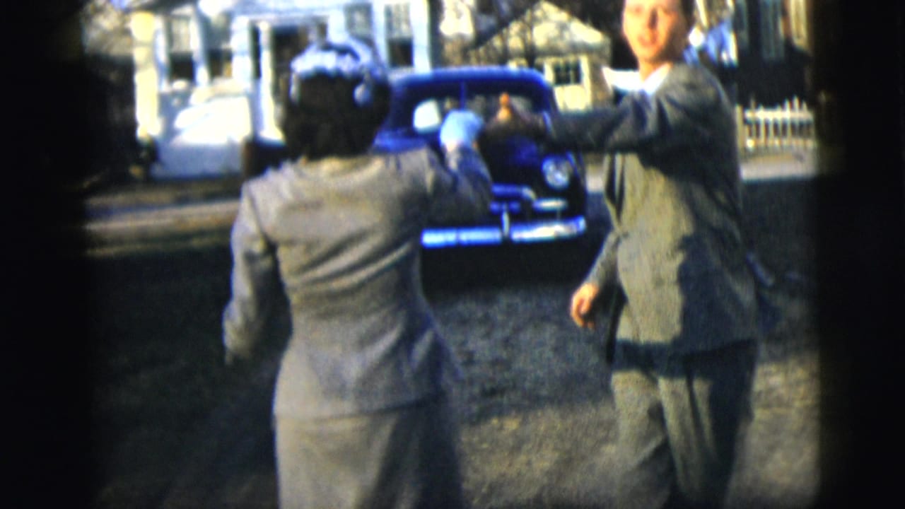A woman in formal clothing faces a suited man on a New York City residential street, with a dark parked car and house fronts in view.
