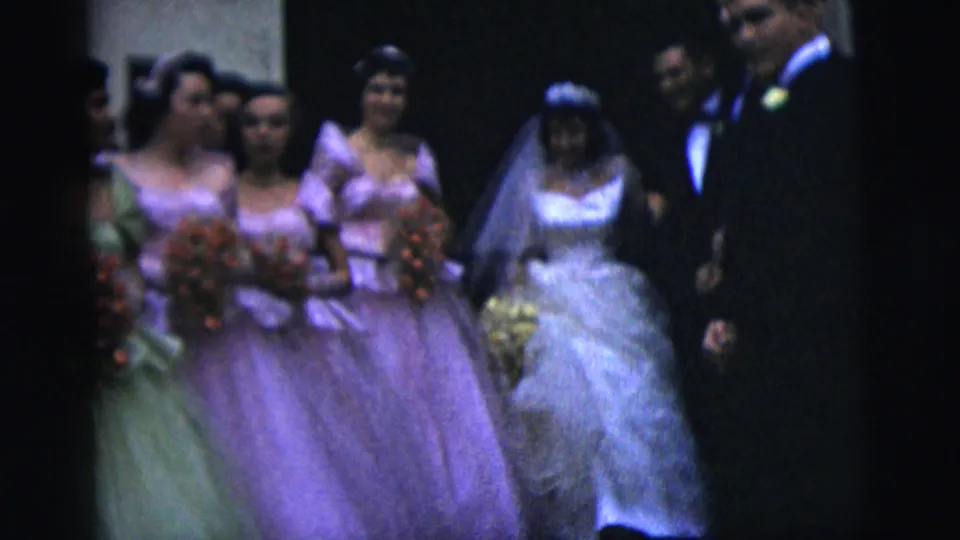 The bride in a veil and white gown walks with bridesmaids in pastel dresses and a suited man, all holding or near floral bouquets.