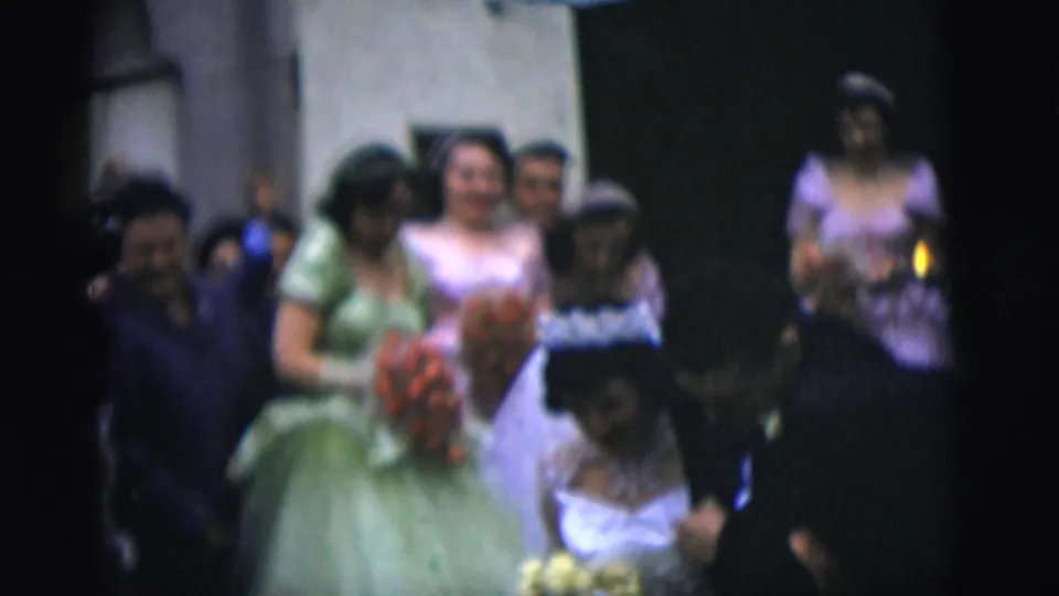 1958 New York City wedding home-movie frame showing bridesmaids, guests, and the bride moving through a dim hallway or hall.