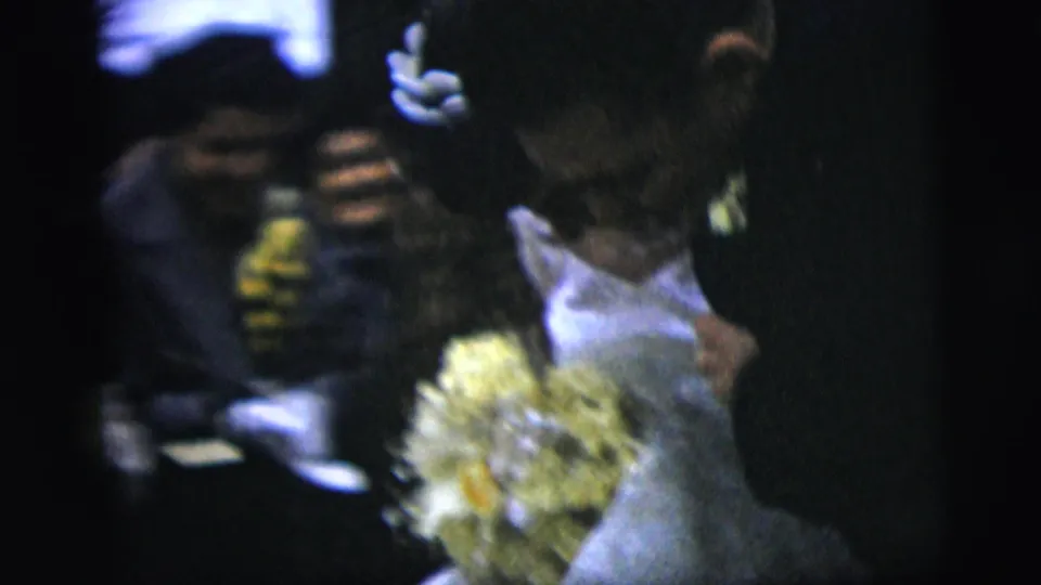 Soft 8mm frame from a 1958 New York City wedding, with the bride, bouquet, and nearby formal guests in close, dark framing.