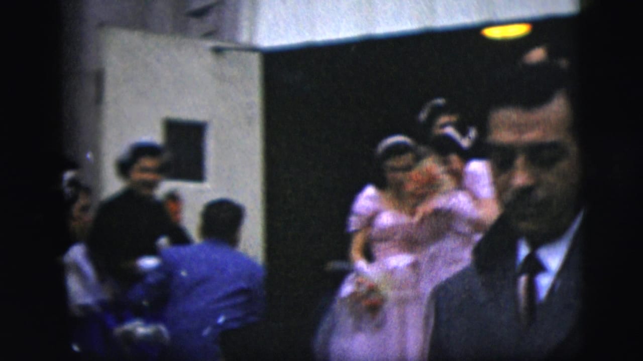 In 1958 New York City home-movie footage, a bride in lavender and a groom in dark formalwear pass through a crowded interior space.