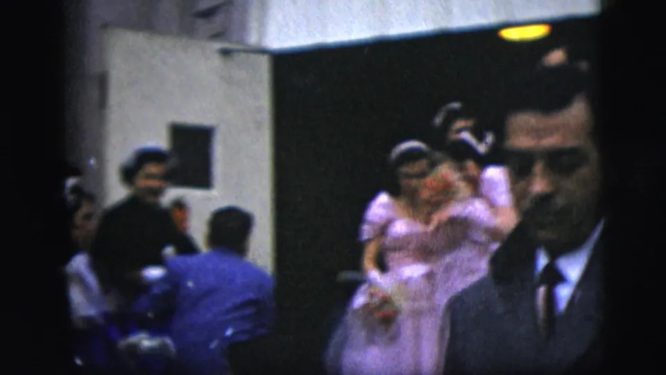 In 1958 New York City home-movie footage, a bride in lavender and a groom in dark formalwear pass through a crowded interior space.