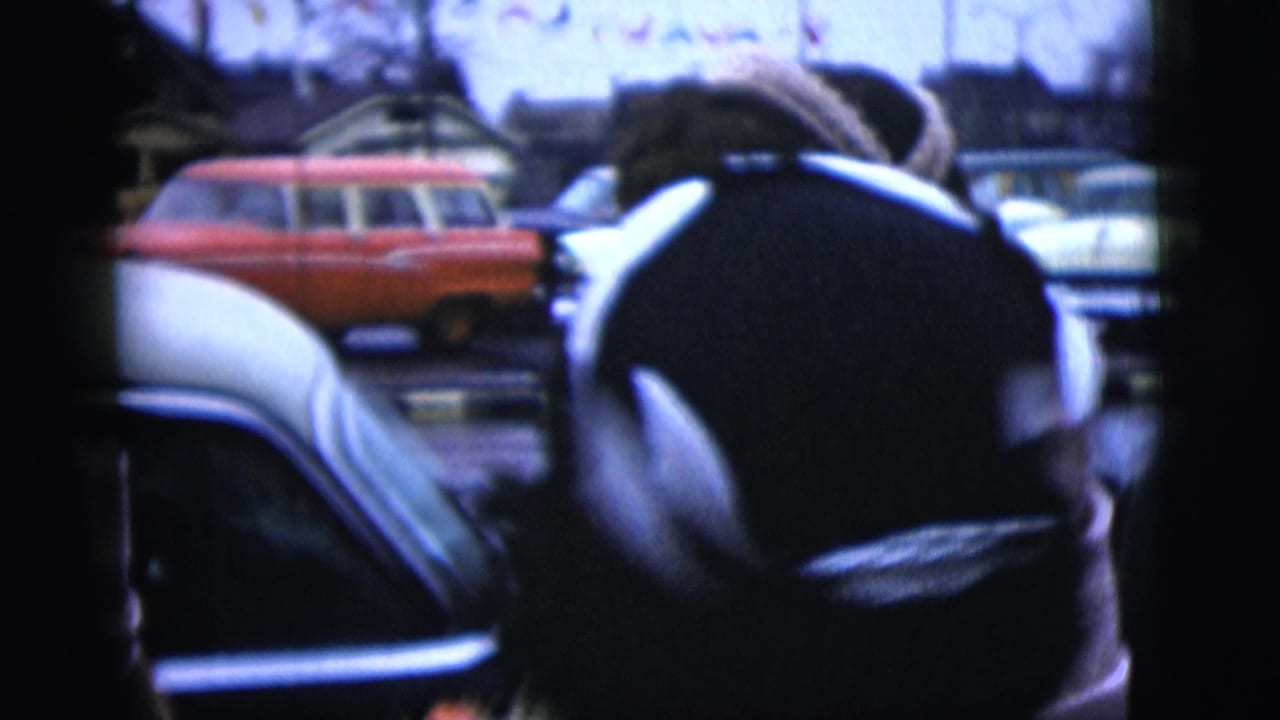 Bundled person seen from behind beside parked mid-century cars on a residential street.