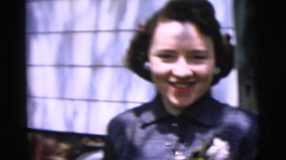 A young woman in 1958 New York City smiles outside, wearing a dark collared dress, pearl earrings, and a light corsage.