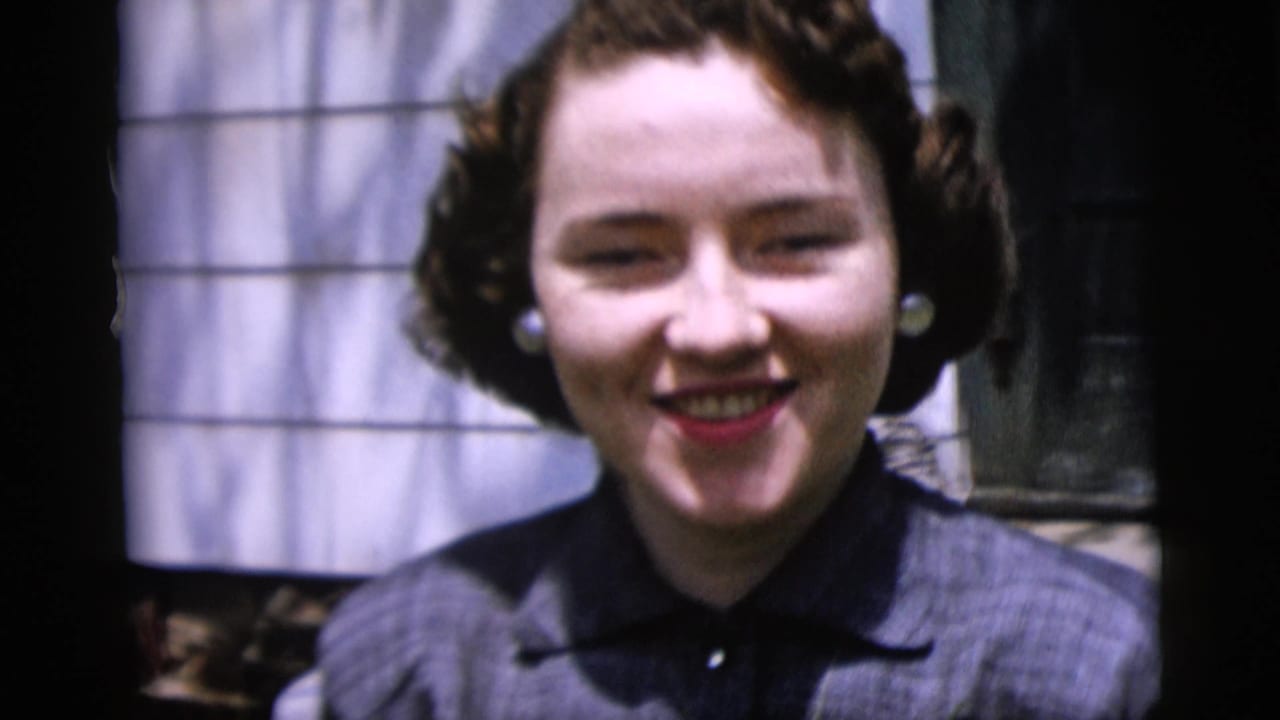 Young woman in a dark dress and pearl earrings smiles toward the camera in 1958 New York home-movie footage.