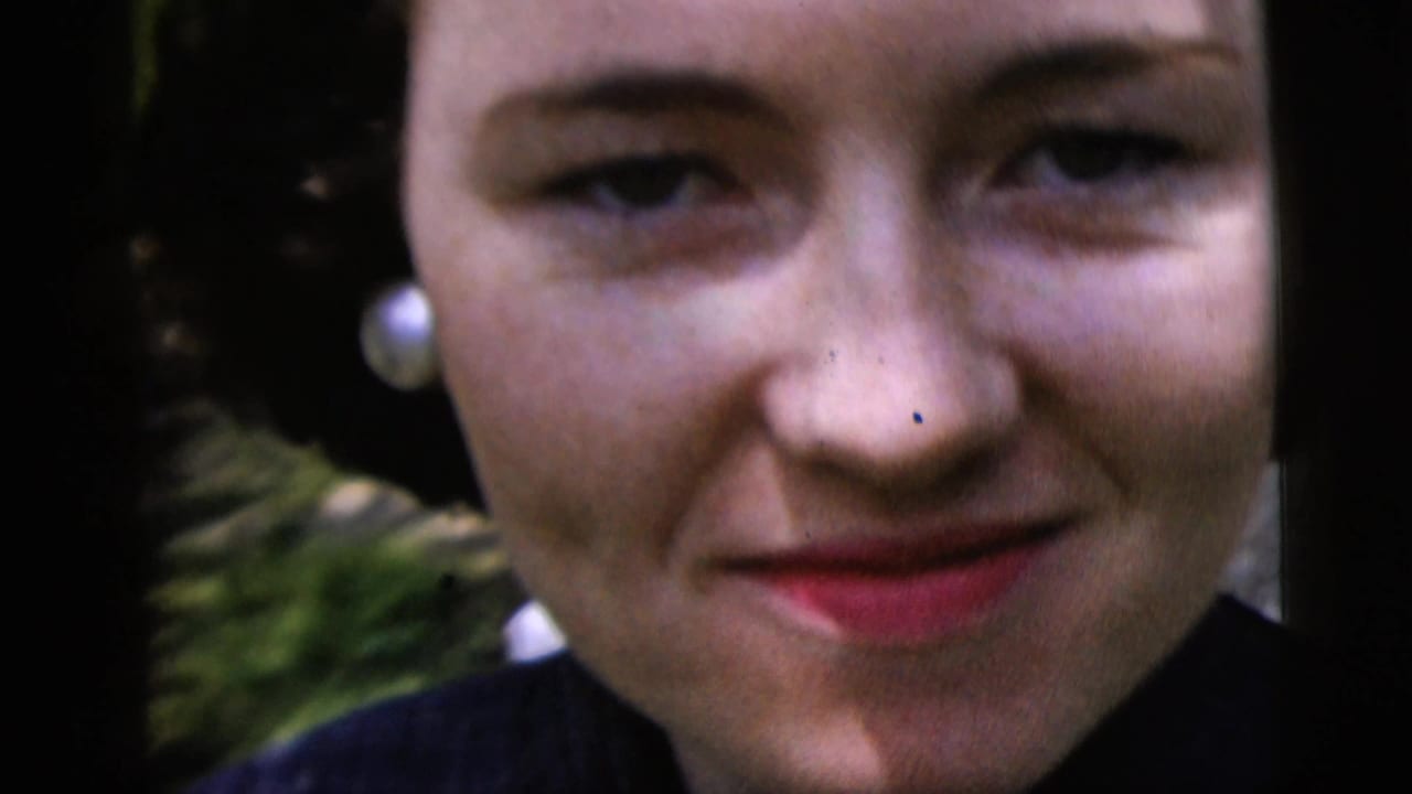 Close-up of the young woman smiling at the camera, with pearl earrings and greenery behind her.
