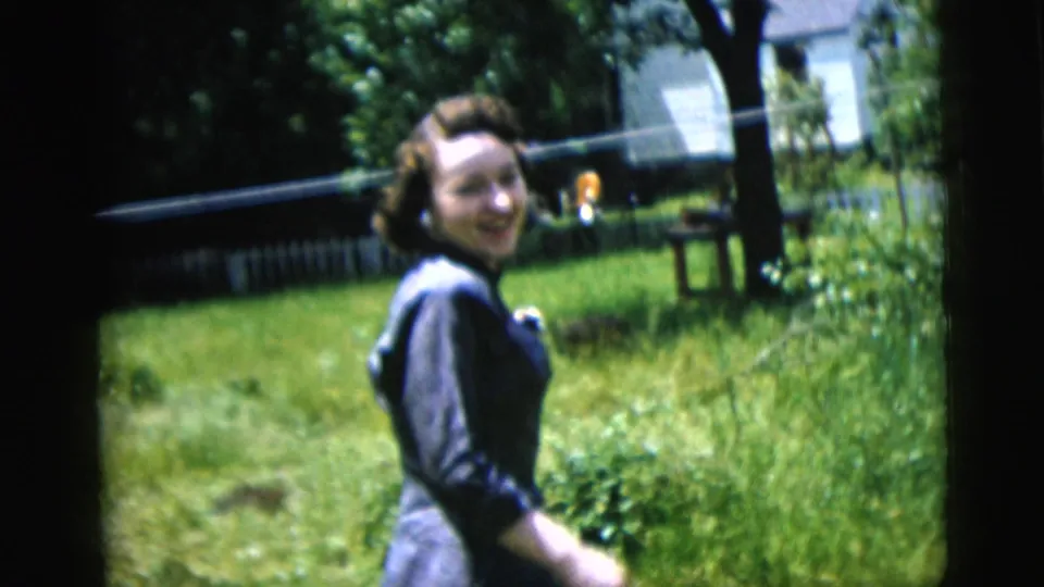 Young woman in a dark dress turns back toward the camera in a sunlit grassy yard with fence and trees.