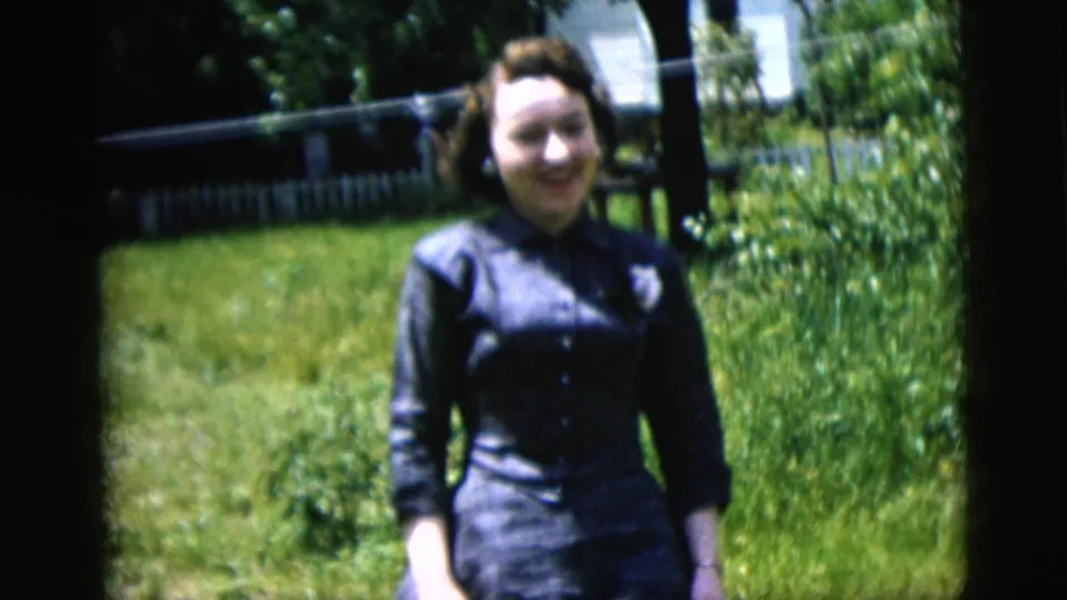 Young woman stands in a sunlit yard, smiling toward the camera with grass, shrubs, trees, and a pale fence behind her.