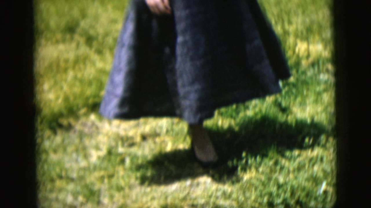 1958 New York City 8mm frame showing the lower half of a woman in a dark blue dress stepping through a grassy yard.