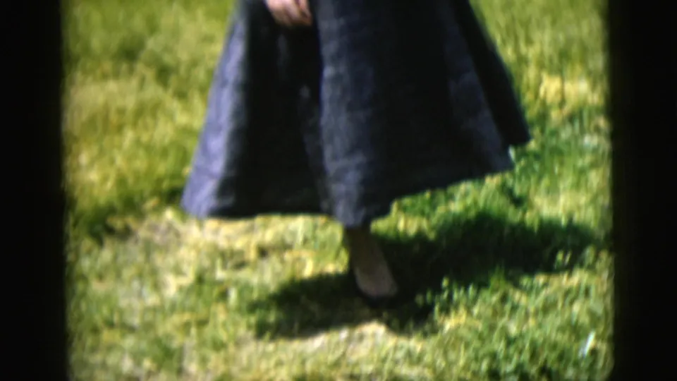 1958 New York City 8mm frame showing the lower half of a woman in a dark blue dress stepping through a grassy yard.