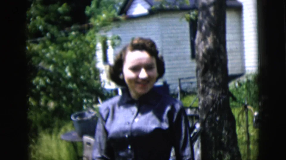 1958 New York City 8mm frame showing a smiling woman in a dark blue dress with a house, tree, and yard behind her.