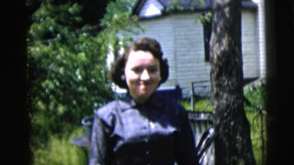 1958 New York City home-movie frame of a woman smiling in a dark blue dress with a tree and house in the background.