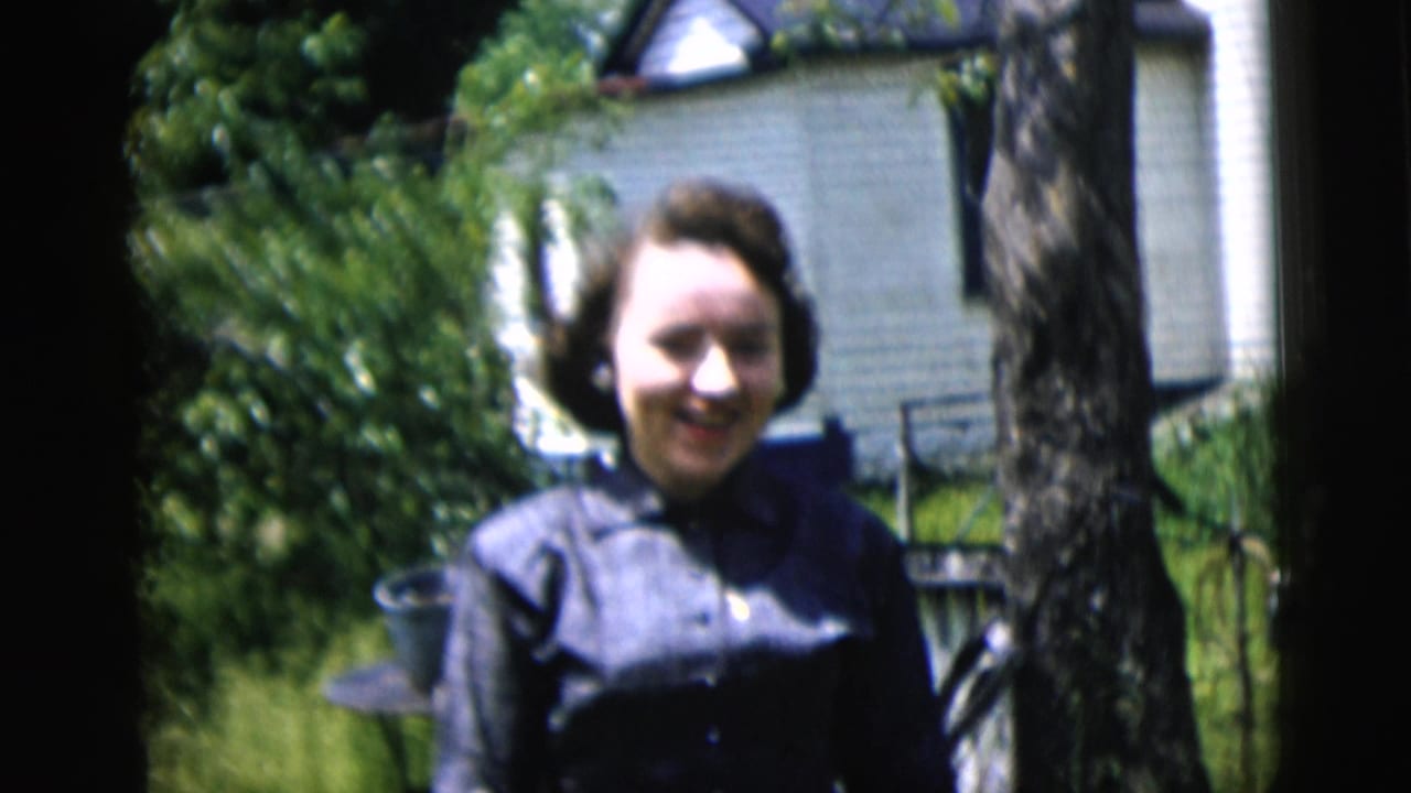 In 1958 New York City home-movie footage, a woman smiles outdoors beside a tree with a white house and greenery behind her.