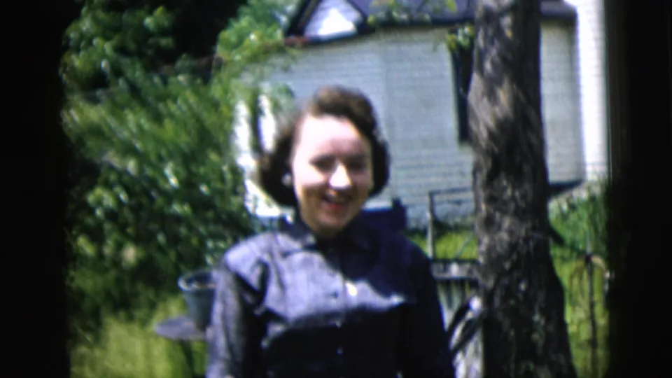In 1958 New York City home-movie footage, a woman smiles outdoors beside a tree with a white house and greenery behind her.