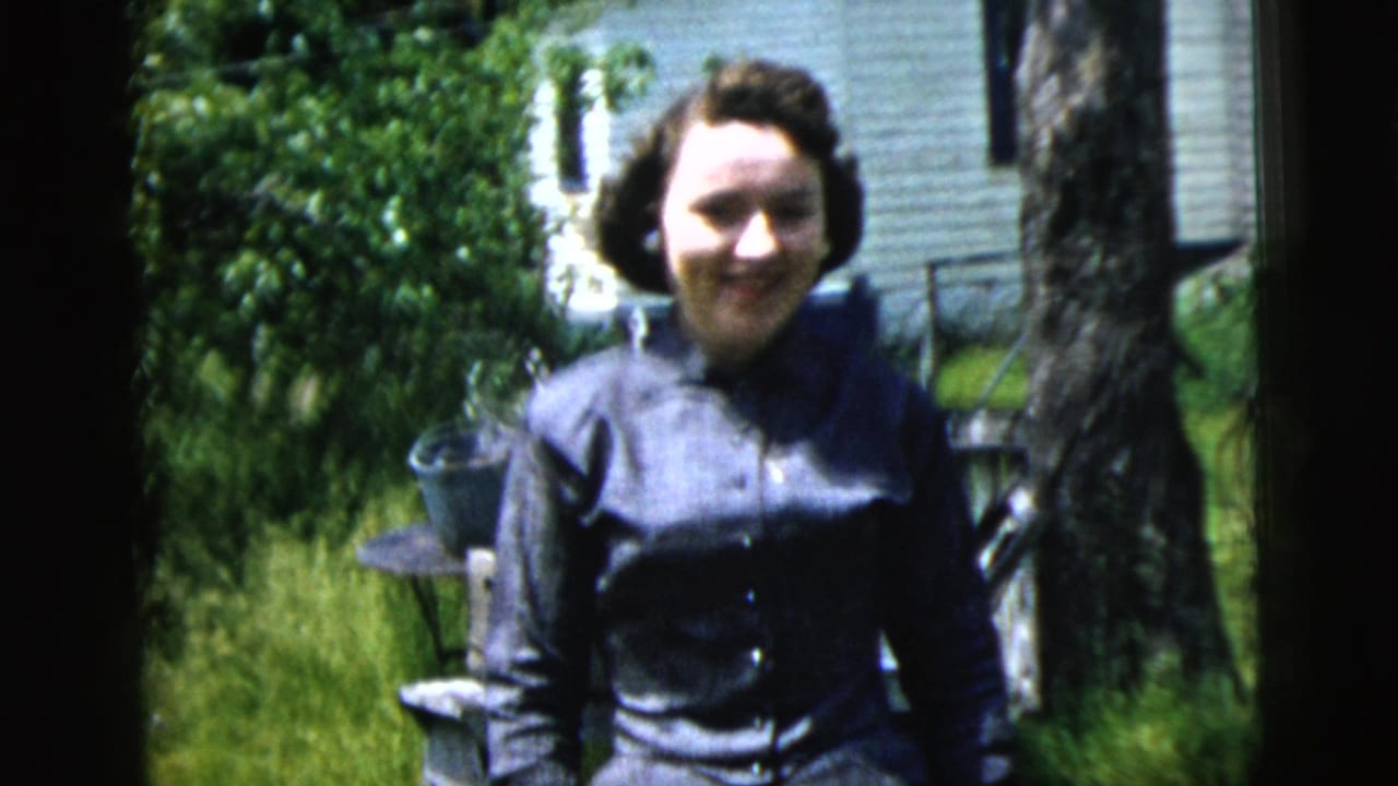 Woman in a dark dress standing outside near a tree and white-sided house.