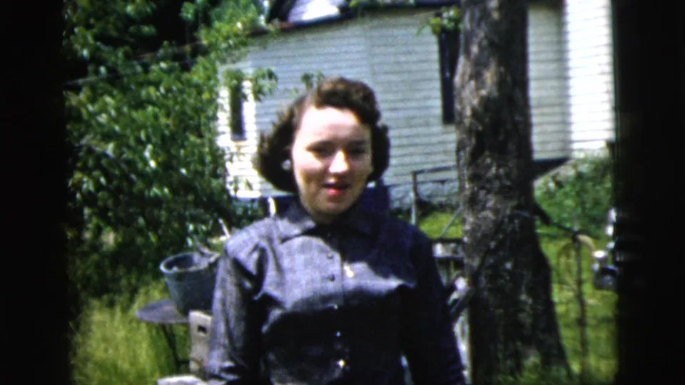 A woman in a dark dress stands in a sunny 1958 New York City residential yard with grass, shrubs, a tree trunk, and house siding behind her.