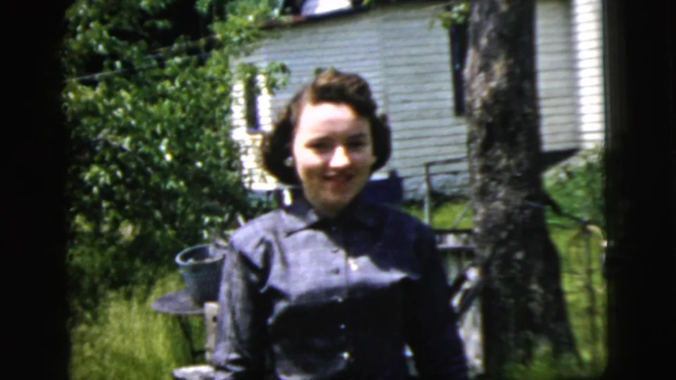 A woman smiles toward the camera in a 1958 New York City backyard, framed by bright grass, leafy plants, a tree, and a white-sided house.