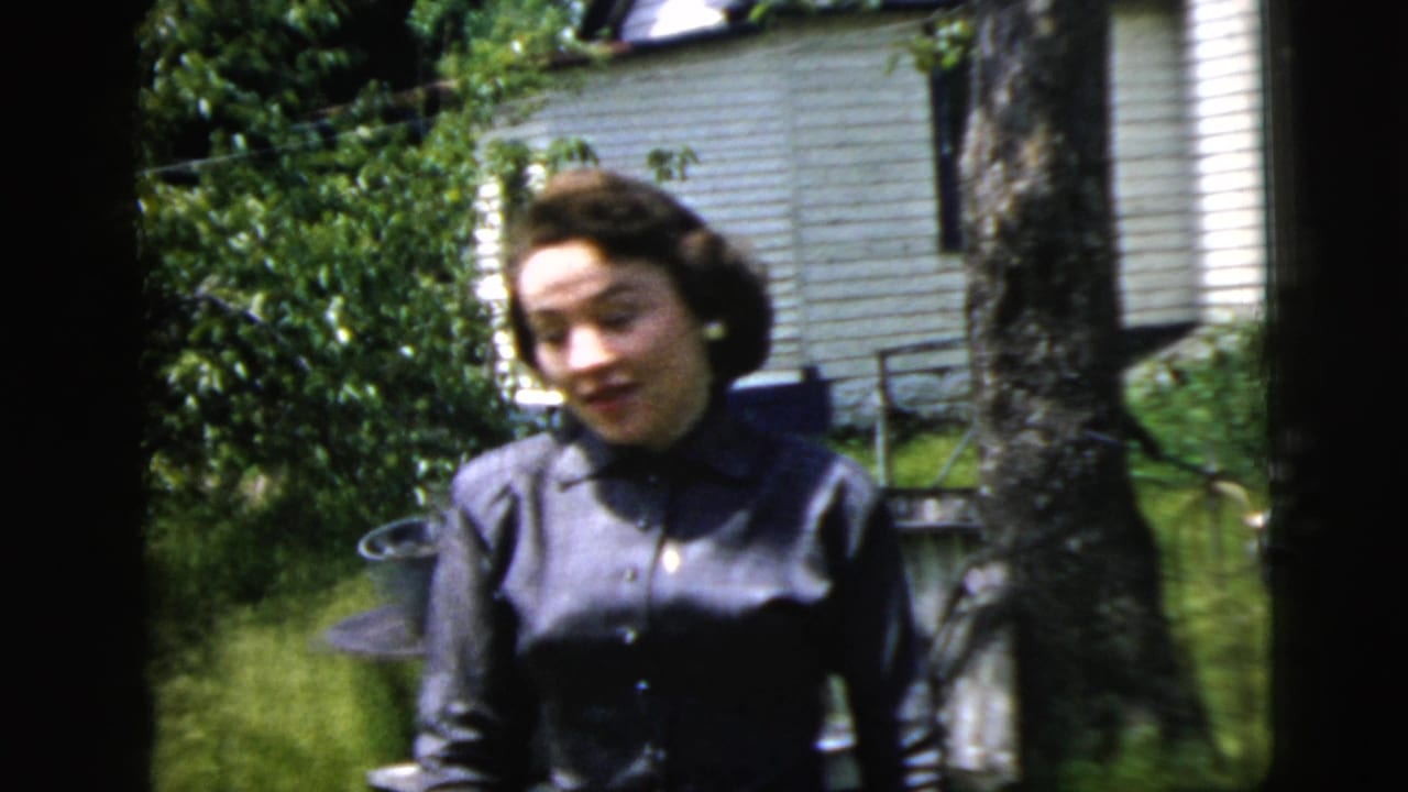 A woman in a blue button-front dress stands in a green garden in 1958 New York City home-movie footage.