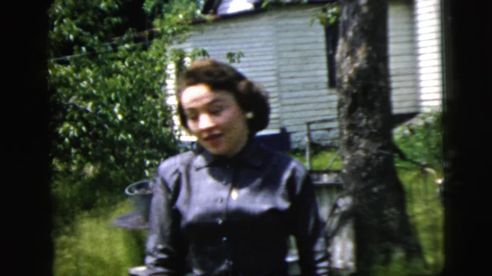 A woman in a blue button-front dress stands in a green garden in 1958 New York City home-movie footage.