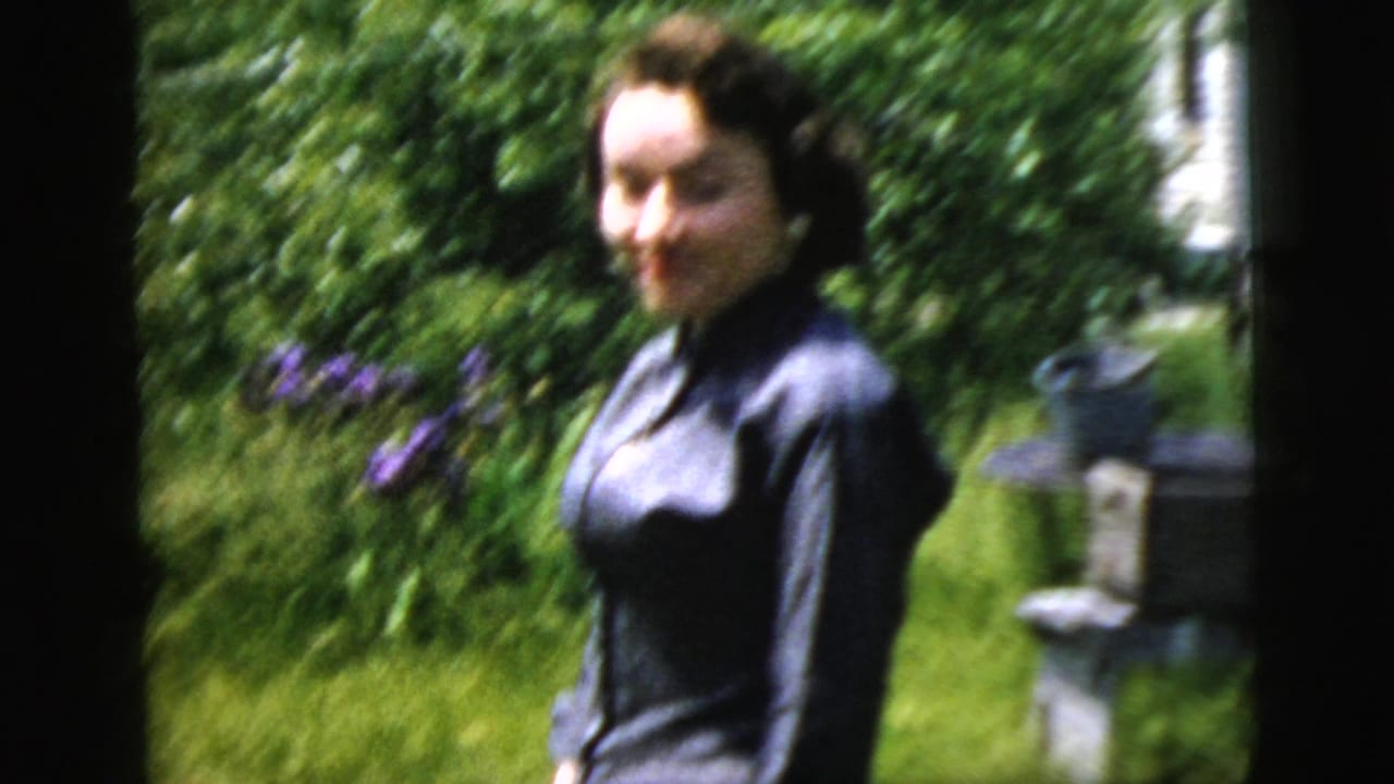 The woman turns slightly in a lush garden, with purple flowers and house siding visible behind her.