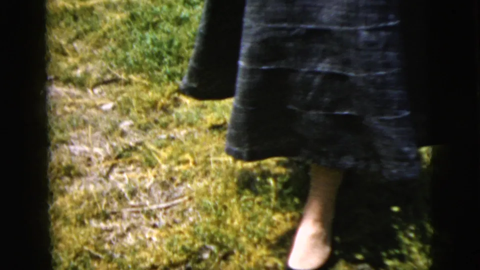 The frame shows the woman's rolled or full skirt, bare legs, and dark shoes standing in uneven garden grass.