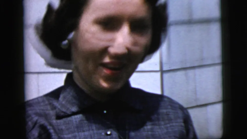 Close-up of the woman smiling toward the camera, with short dark hair, earrings, and white exterior siding behind her.