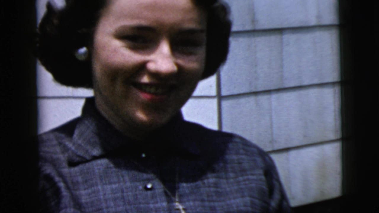 Young woman in a dark collared dress smiles toward the camera in 1958 New York home-movie footage.