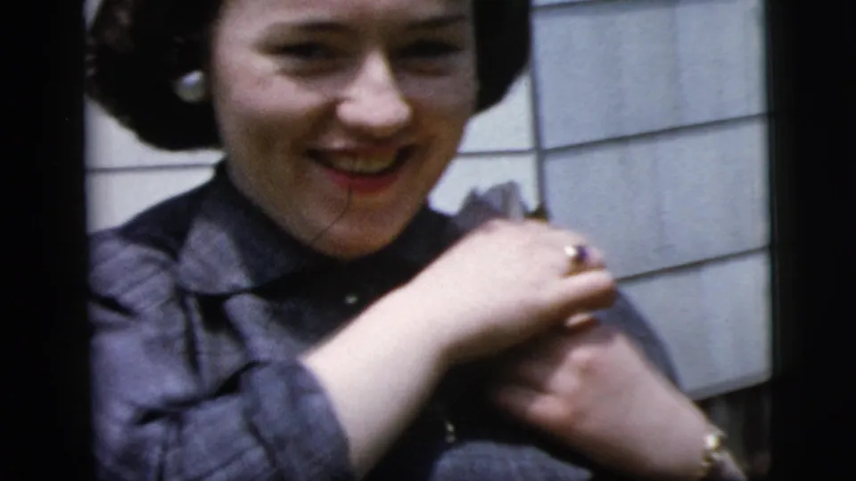 In 1958 New York footage, the woman smiles as she raises her hands to pin or adjust a corsage on her shoulder.