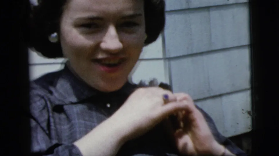 Close 8mm frame of a woman in a dark dress holding a corsage near her left shoulder in a New York home movie from 1958.