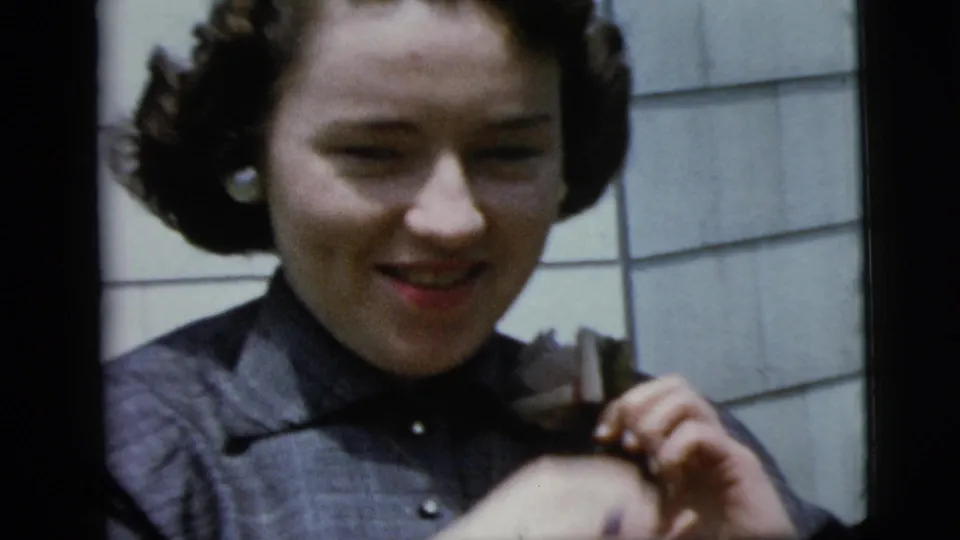 The woman looks down slightly while adjusting the corsage on her dark dress in 1958 New York home-movie footage.