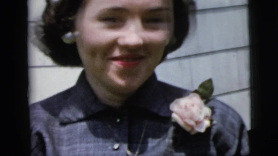1958 New York City home-movie frame of a young woman smiling in pearl earrings, dark blouse, and pale rose corsage outdoors.