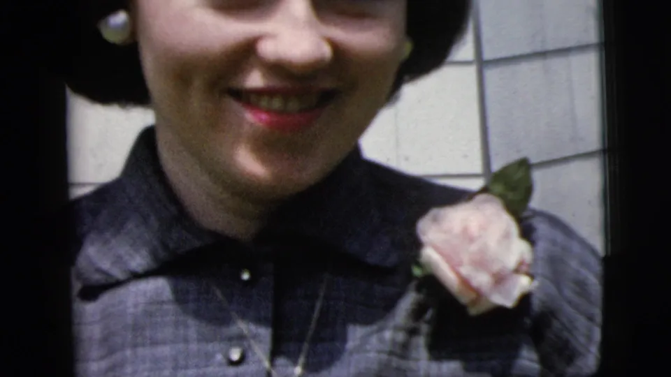 Close 1958 New York City 8mm view of the smiling woman with pearl earrings, red lipstick, dark blouse, and rose corsage.