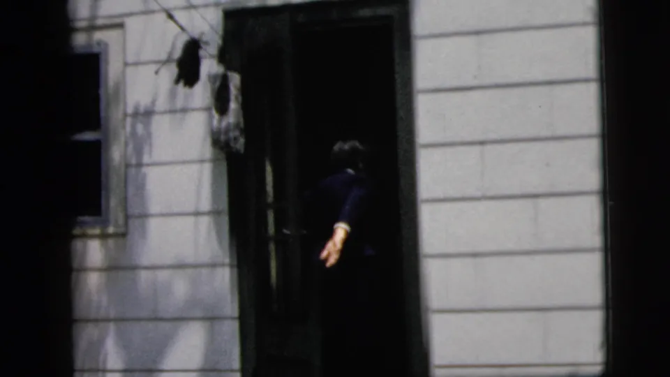 1958 New York City 8mm frame showing the woman from behind near a dark doorway on a white-sided house exterior.