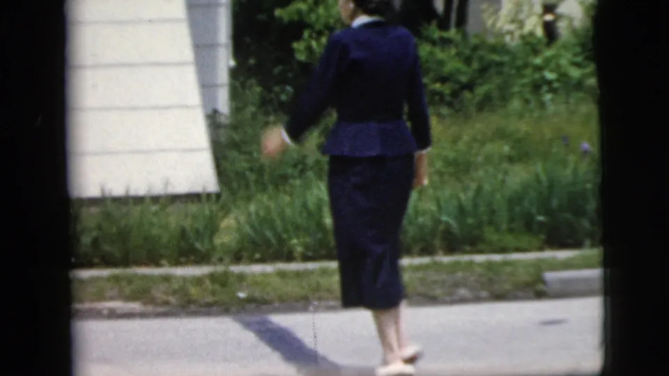 Rear view of a woman in dark blue mid-century clothing walking beside a grass verge and light house siding in New York home-movie footage.