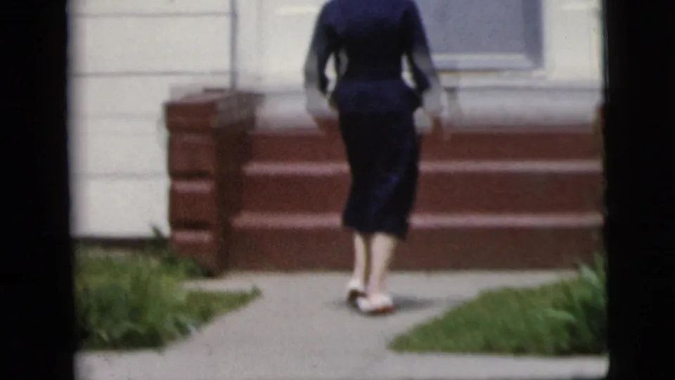 Woman in a dark blue outfit approaches red brick steps and a light-sided residential exterior in 1958 New York home-movie footage.