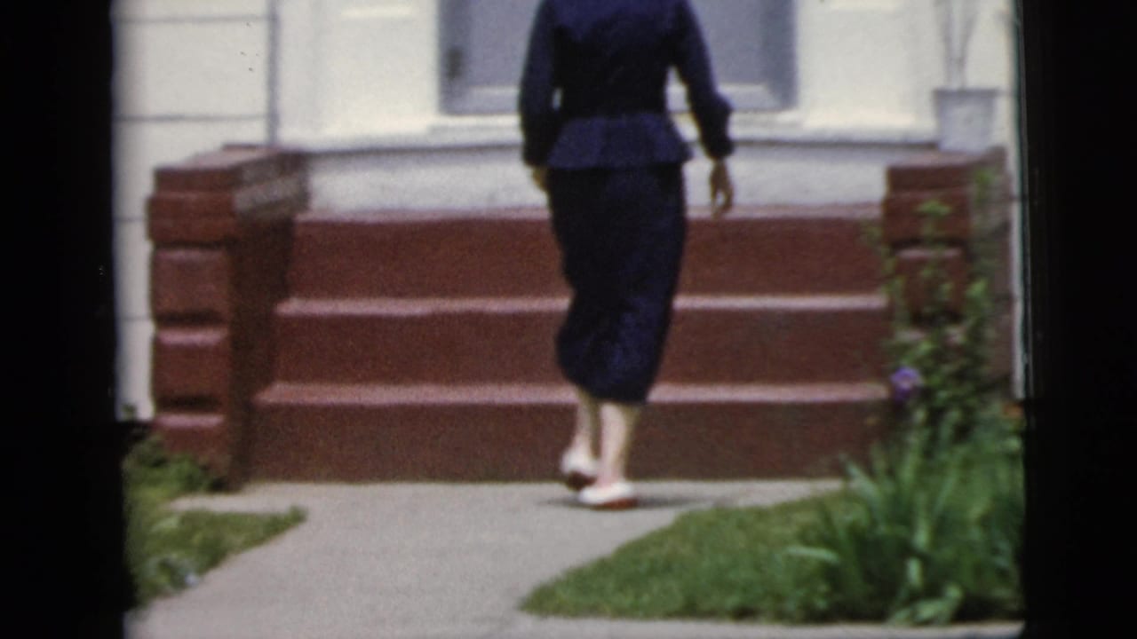In 1958 New York City home-movie footage, a woman in dark clothing walks up red steps toward a white residential doorway.