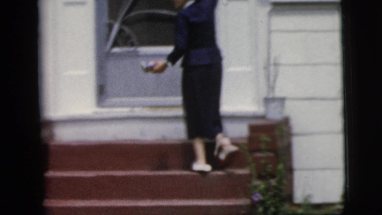 Woman steps toward a gray door while holding something in one hand.